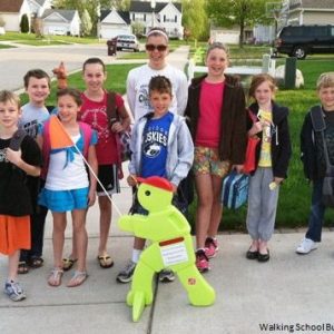 Bunch of students waiting to cross the street with yellow doll with orange flag 