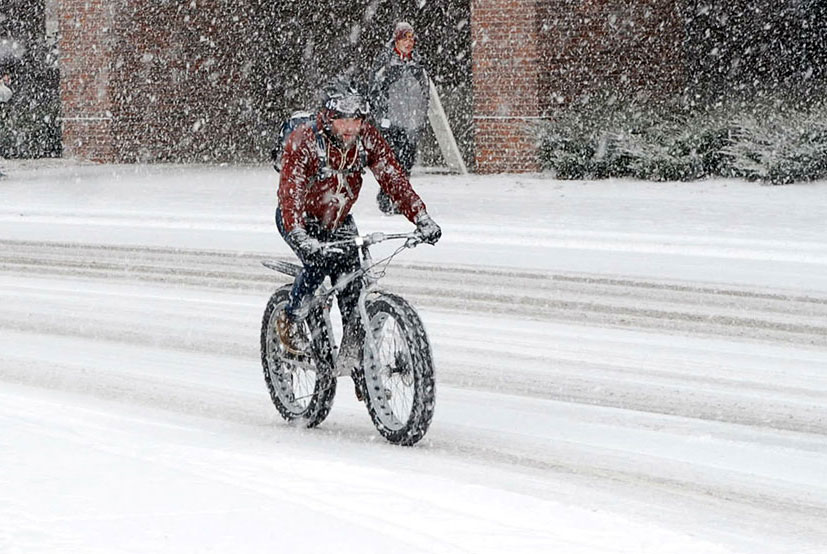 Person on a fat bike biking down a snow covered road with a person walking in the background.