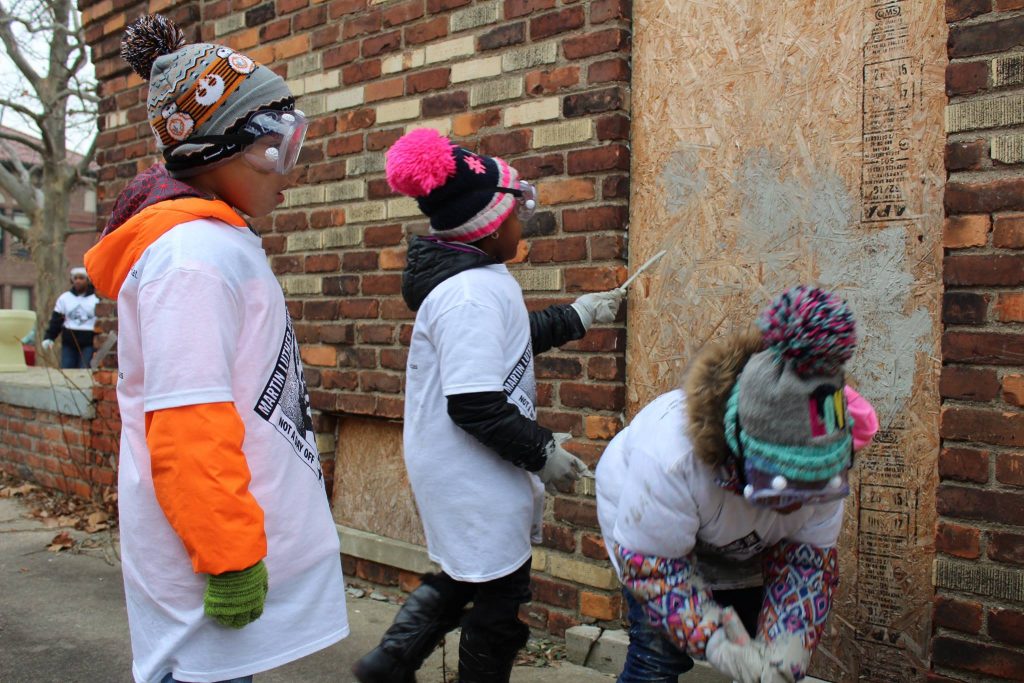 Young kids in hats, googles, and work gloves paint boarded up buildings along a Safe Routes corridor in Detroit as part of MLK Service Day.