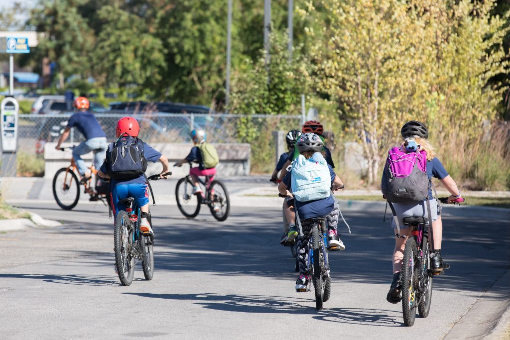 Image of a handful of students biking away from the camera with an adult leading them as part of a bike train in Traverse City. Background is of trees. and a parking lot.