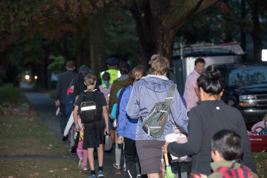 A few of a large group of students and parents walking down a sidewalk while in the dark hours of the morning as part of a Walk to School Day event.