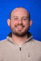 White man with facial hair wearing camel pullover with deep blue background