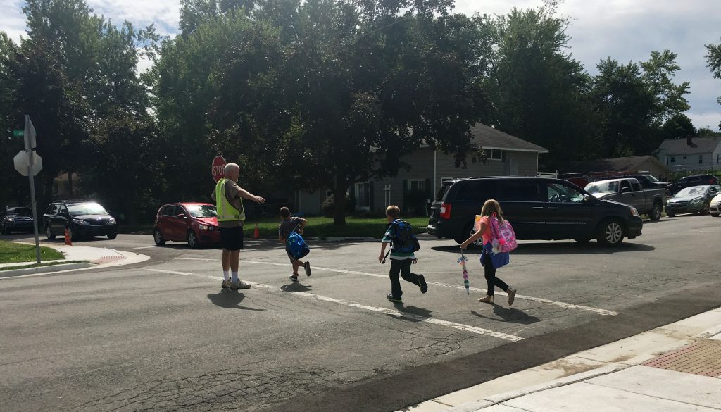 three students run across a three way intersection with the help of a crossing guard where there were SRTS infrastructure improvements made.