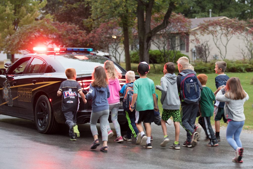 a group of students walk behind a police car that is holding traffic for them to participate in Walk to School Day in an area with limited infrastructure for safe walking.