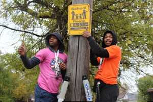 Two students wearing pink and orange t-shirt pointing to the yellow sign on the tree 