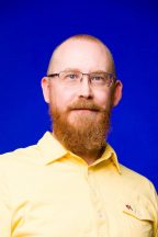 White Man with glasses and orange facial hair in yellow collared shirt with deep blue background