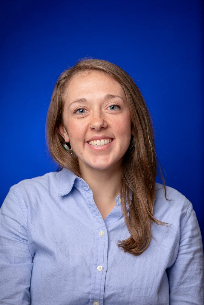 White woman with long brown hair wearing blue collared shirt with deep blue background