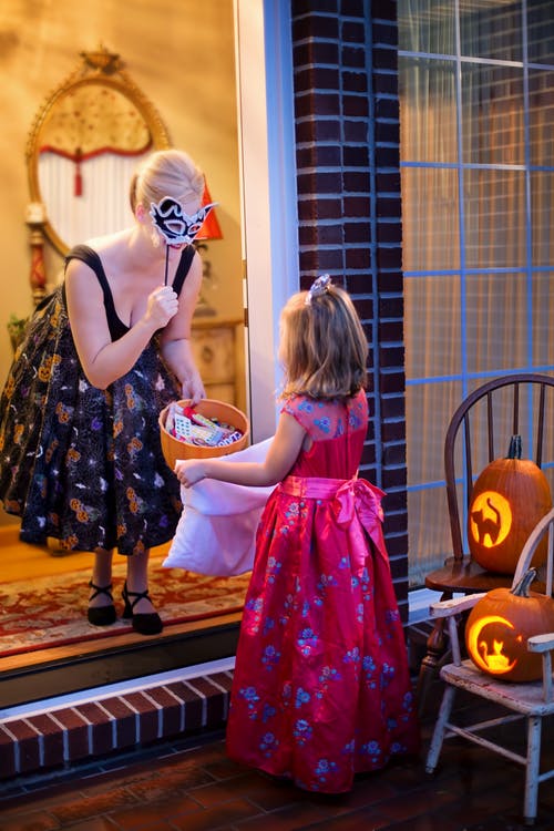 girl in a pick costume at a doorway hold trick or treat bag out to adult holding up a mask and offering a bowl of candy