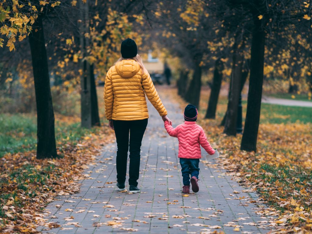 a woman and her child hold hands and walk down a foliage and tree lined pathway of pavers.