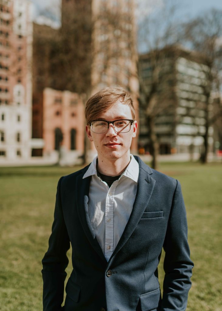 White man with light hair, wearing glasses and suit jacket with park and buildings in the background.