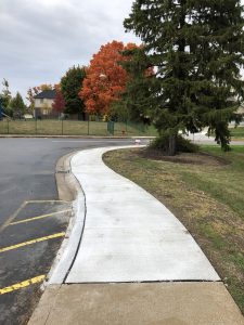 Sidewalk agjacent to school parking lot with yellow parking lot paint alongside the sidewalk and fall trees in the background. 