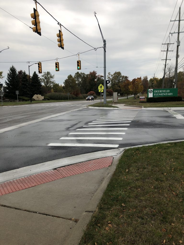 Intersection crossing at light with new ADA ramps, and high vis yellow school crossing sign. School build sign visible in the distance.