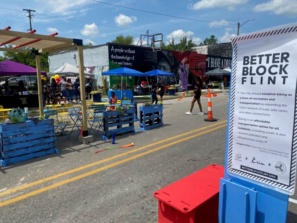 A brightly painted parklet is set up in a closed road area as part of the Better Block Flint event. Poster for the event seen in the foreground.