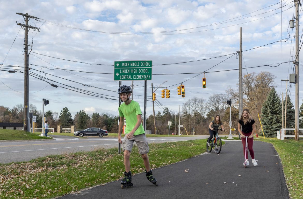 Three student use a multi use path by roller skating, scootering, and biking as they move away from an intersection.