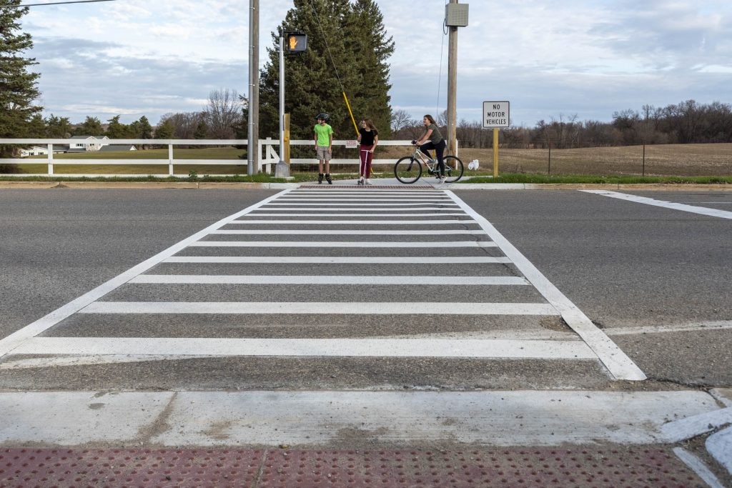 Student roller skater, scooter, and biker are on the other side of an intersection with pedestrian signal preparing to cross.