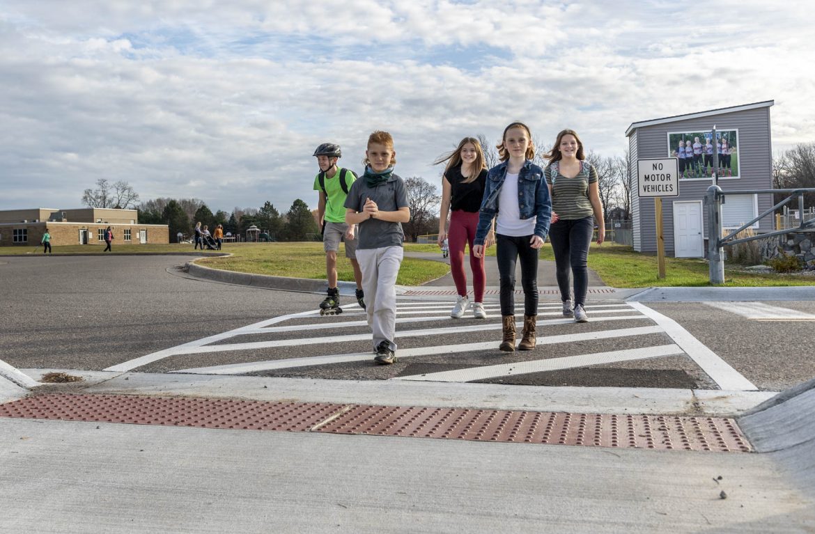 Five students walking and roller skating across a marked cross walk with the school in the background.
