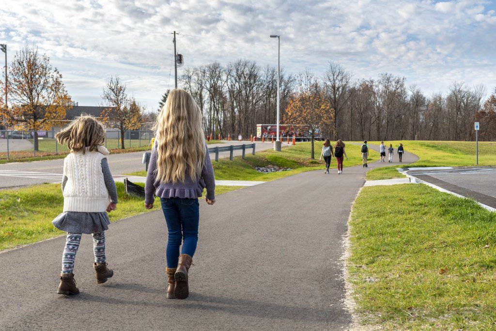 Students are walking with their back in viewon a trail positioned between a parking lot and road. the two closest students are younger girls in the left near corner.