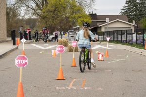Portland Middle School Bike Rodeo event showing student on lime green bike riding away through an obstacle to practice stopping accurately. Other students on bikes and adult volunteers in the background.