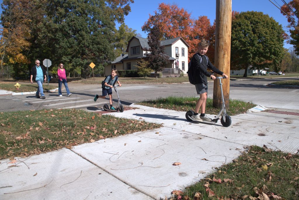 Two children scooter onto new sidewalks, while adults walk behind utilizing crosswalk. Photo Credit MDOT