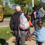A student bikes on a sidewalk with their father walking beside them. Other parents and children walk and bike ahead of them. A school bus passes on the road beside the sidewalk. MDOT photo credit. 