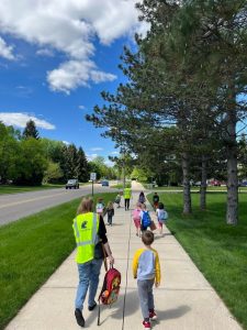Students with adult volunteers walking along sidewalk with roadway and trees alongside them