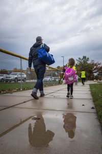 Adult with a bag and student walk to school with puddles on the foreground and overcast storm clouds in the sky in the background.