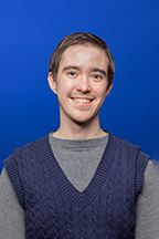 Person with brown hair and brown eyes smiling wearing a blue sweater vest over a grey shirt with a deep blue background