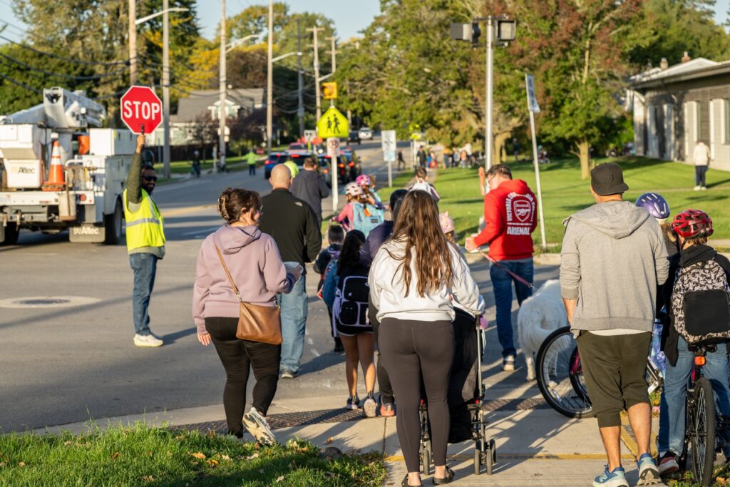 Marble Elementary students and parents walk and bike across an intersection while a crossing guard holds aloft a stop sign to control vehicle traffic. MDOT Photo Credit