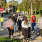 Marble Elementary students and parents walk and bike across an intersection while a crossing guard holds aloft a stop sign to control vehicle traffic. MDOT Photo Credit
