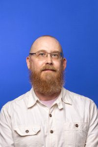 White man with wire glasses and orange facial hair in white collared shirt with blue background