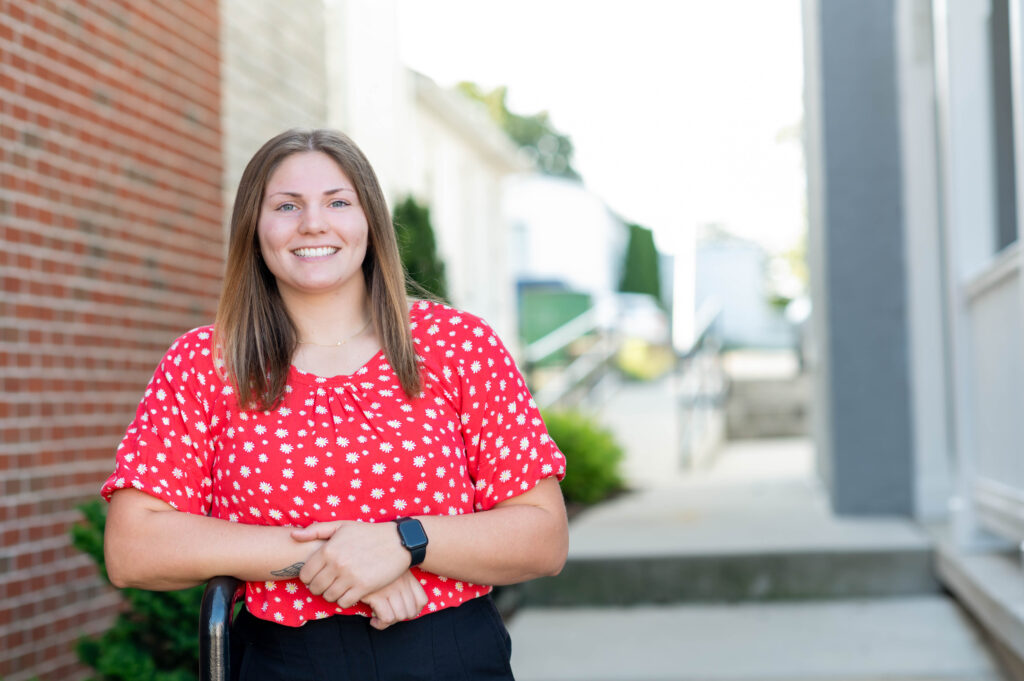 White worman with long brown hair wearing a red and white floral patterned shirt stands in front of a blurred background.
