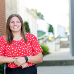 White worman with long brown hair wearing a red and white floral patterned shirt stands in front of a blurred background.