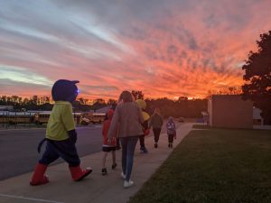 Students walk with mascots toward school during the sunrise.