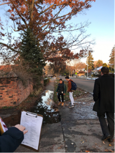 Section of a walking audit where there is a puddle covering the sidewalk due to poor drainage. Two students are standing by the puddle with a man approaching. A clip board with the audit form is in the foreground. The background is a residential street with fall trees.
