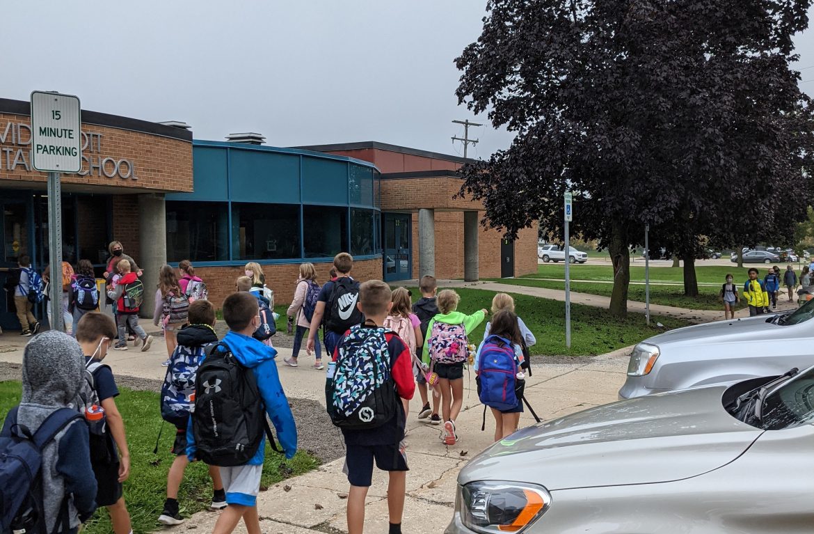 students with backpacks walking toward the entrance of a school with park parking along sidewalk towards building