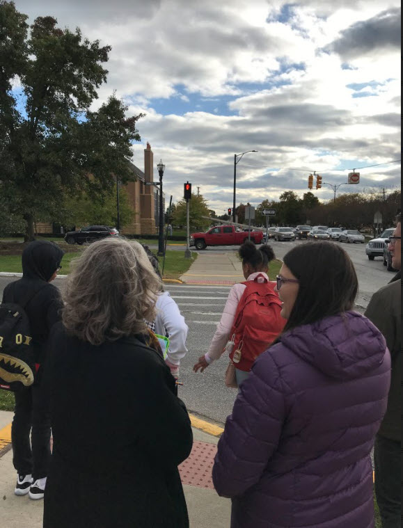 Adults and students participating in a walking audit. Group is facing away and approaching a crosswalk. The sky is full of clouds.