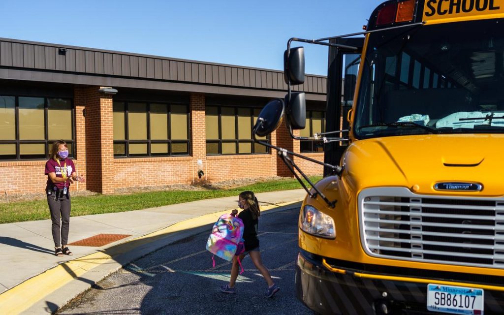 student exits school bus at school building with masked staff welcoming them.