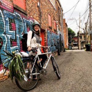Female with helmet and glasses beside bike with a plant on its back rack with graffiti art on brick buildings in the background