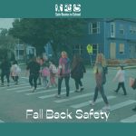 Students and parents walk along a zebra stripped street crossing in the dim of morning with car lights shining on them. Text is the Safe Routes to School logo on top and "Fall Back Safety" at the bottom.