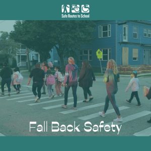 Students and parents walk along a zebra stripped street crossing in the dim of morning with car lights shining on them. Text is the Safe Routes to School logo on top and "Fall Back Safety" at the bottom.