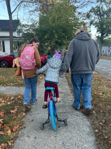 Two adults walking accompany one child biking on sidewalk.