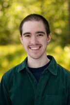 Person with brown hair and brown eyes smiling wearing a green button down shirt with trees in the background