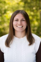 White woman with brown hair and brown eyes wearing a brown shirt with a white sweater vest over it with trees in the background