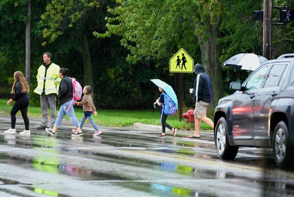 Students, some with and without umbrellas, cross at crosswalk with crossing guard on a raining morning at Sycamore Elementary