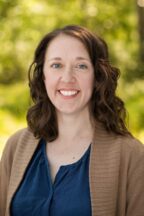 White woman with wavy brown hair and blue eyes wearing a blue top and brown cardigan with trees in the background