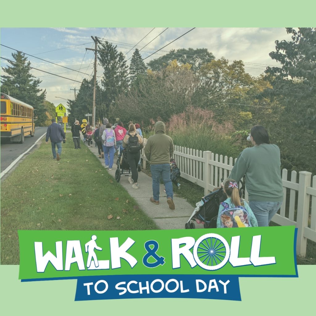 Walking school bus of adults and students walking along sidewalk on cooridor with school bus driving in roadway and iwth school crossing signage in the distance.